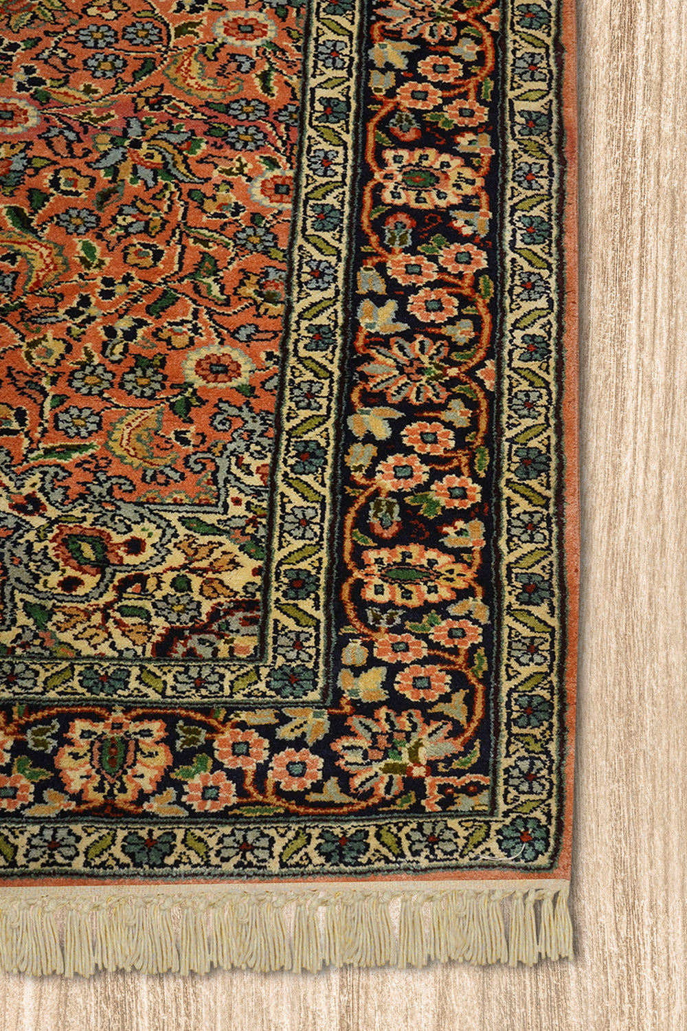 A close-up of the corner of a Kirman Persian rug, highlighting its intricate floral border design and delicate fringe. The rich peach and navy hues contrast beautifully with the finely detailed green and ivory patterns. The rug is placed on a light wooden floor, emphasizing its elegant craftsmanship.
