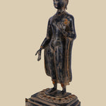 Brass standing Buddha figure in the Gandhara style, with flowing robes, one hand extended, and a serene expression, atop a detailed lotus base.